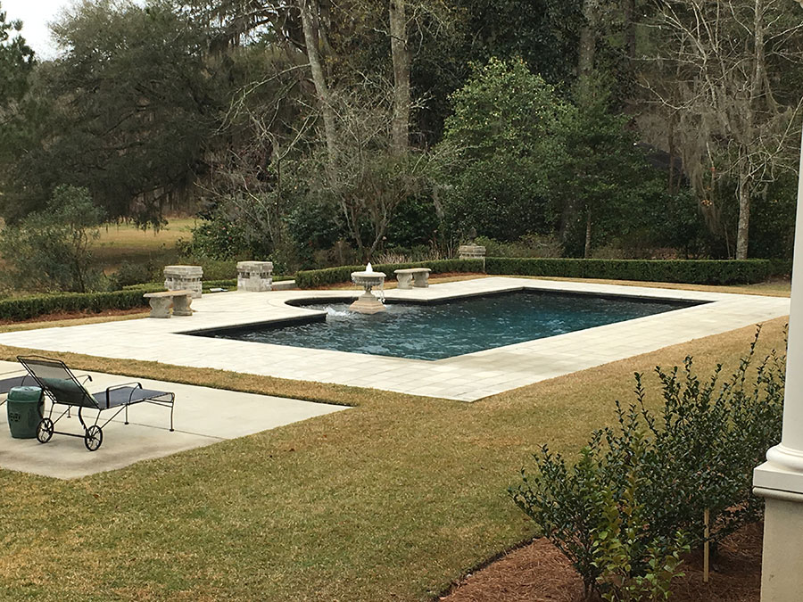 Mystic Pools custom saltwater gunite pool in Tallahassee with stone paver decking, classical tiered fountain, stone pedestal seating, and a manicured boxwood hedge surrounded by wooded landscaping