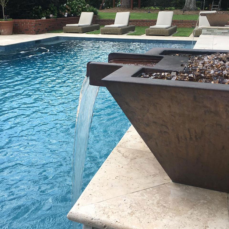 Close-up of Mystic Pools custom gunite pool in Thomasville featuring a concrete fire and water bowl with glass rock media cascading a sheer spillway into the pool, with travertine coping and lounge chairs in the background