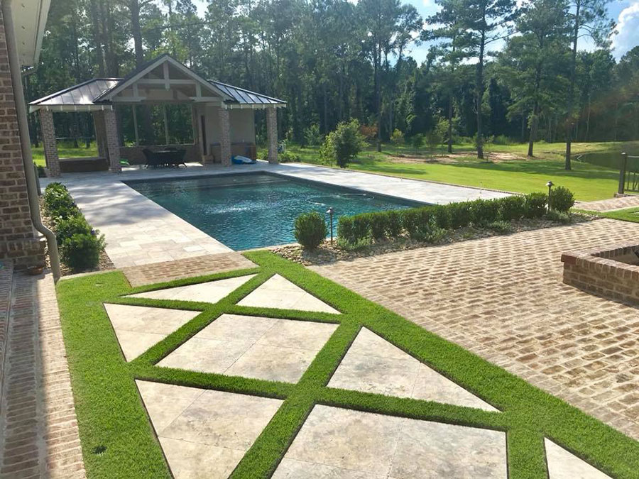 Mystic Pools pool renovation in Thomasville featuring a rectangular pool with travertine decking, geometric paver and grass patio pattern, decorative hedge border, brick paver patio, and an open-air pavilion with fire pit