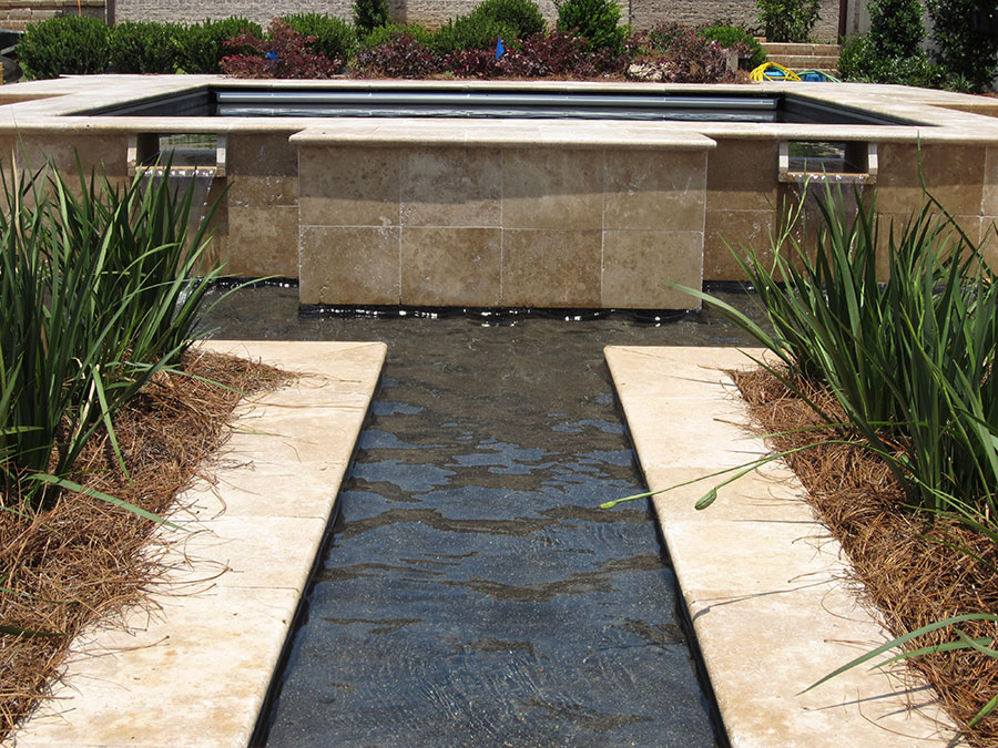 Mystic Pools remodeled pool feature in Thomasville with raised travertine-clad spa wall, automatic pool cover, dual scupper spillways, and a narrow linear water channel flanked by ornamental grasses and pine straw landscaping