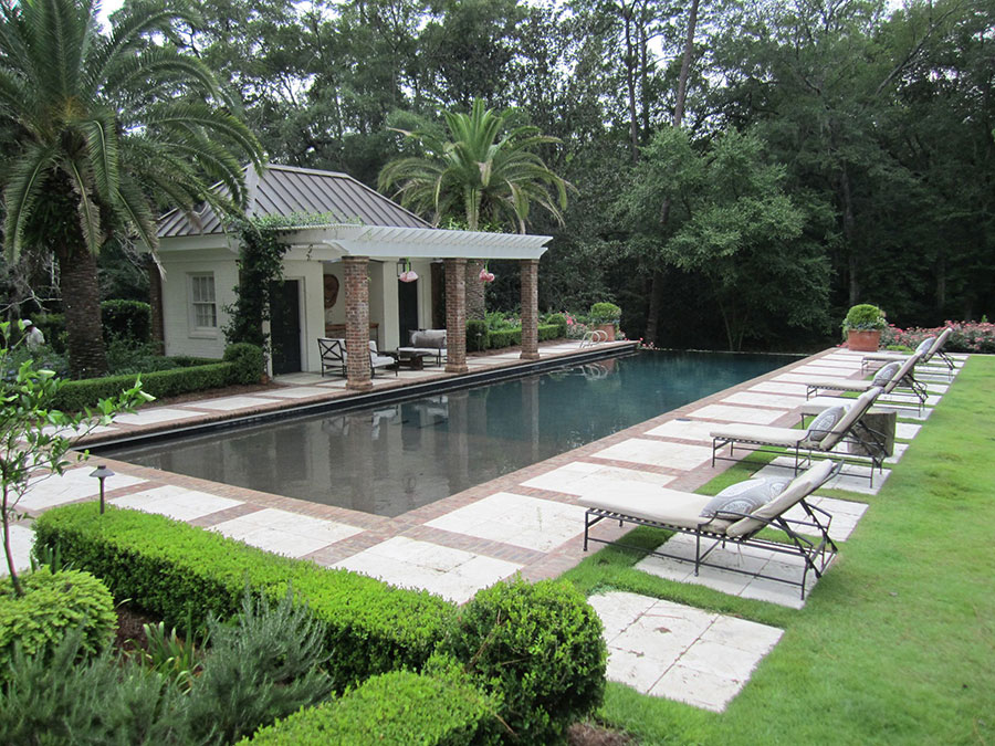 Mystic Pools swimming pool construction in Thomasville featuring a formal dark-finish rectangular pool, brick and travertine paver walkway, padded lounge chaises, manicured boxwood hedges, and a brick pool house with a vine-covered pergola