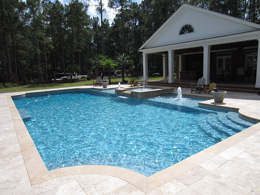 Mystic Pools custom rectangular gunite pool in Tifton with attached spa, dual fountain jets, travertine paver decking, Adirondack seating, and a classic Southern pool house with columned porch surrounded by tall pines
