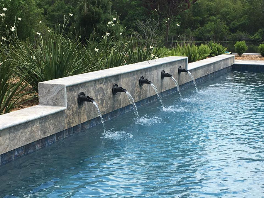 Mystic Pools custom gunite pool construction in Tifton featuring a raised travertine wall with five bronze scupper spouts cascading water into the pool, surrounded by ornamental grasses and flowering plants