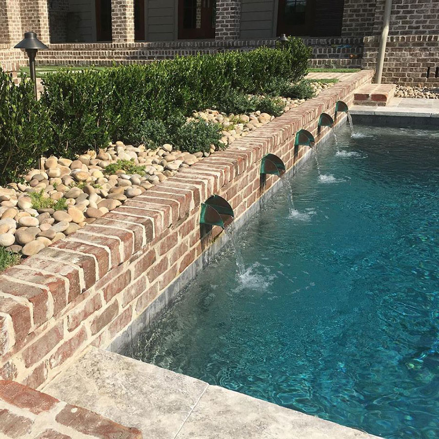Mystic Pools pool renovation in Tifton featuring a brick retaining wall with five bronze scupper spillways, river rock landscaping beds with boxwood hedges, and a brick estate home in the background
