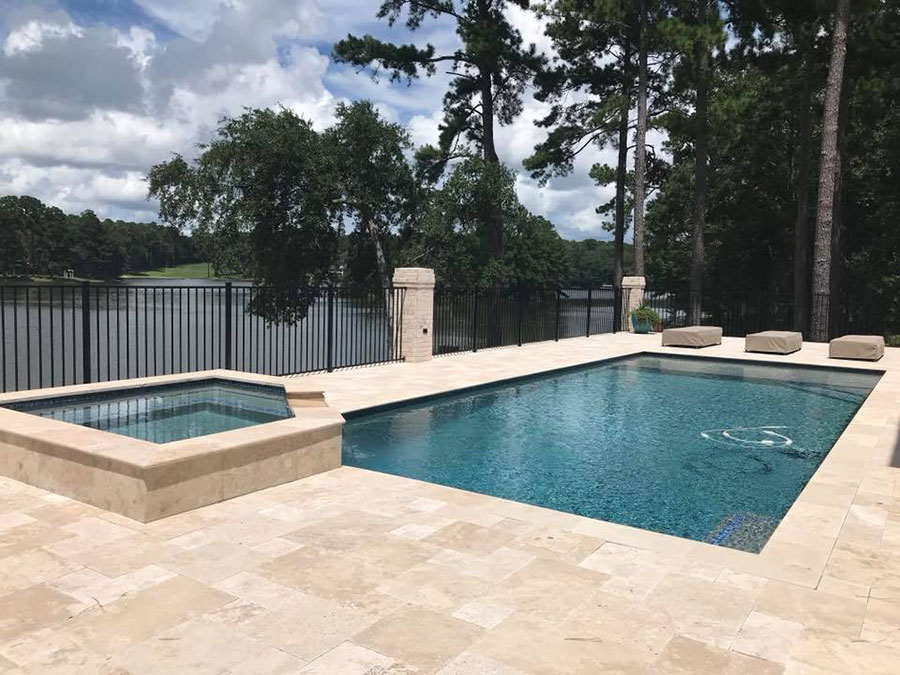 Mystic Pools custom saltwater gunite pool in Tifton with raised square spa, travertine paver decking, lounge ottomans, wrought iron fencing, and a scenic lakefront view framed by tall pine trees