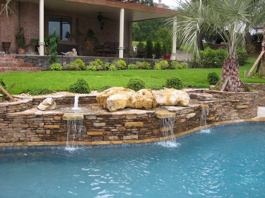 Mystic Pools custom gunite pool construction in Valdosta featuring a curved stacked stone grotto wall with natural boulder accents and multiple cascading waterfall spillways into the pool