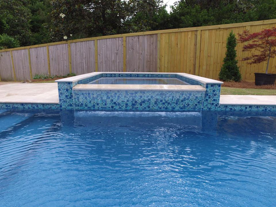 Mystic Pools pool renovation in Valdosta featuring a raised square spa with blue and green mosaic glass tile, travertine coping, and a wood privacy fence with landscaped backyard