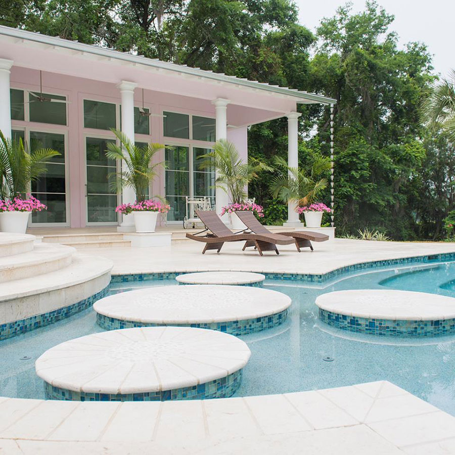 Mystic Pools remodeled freeform gunite pool in Valdosta featuring circular travertine stepping stones, blue mosaic tile accents, lounge chairs, colorful flower planters, and a pink columned estate home with tropical landscaping