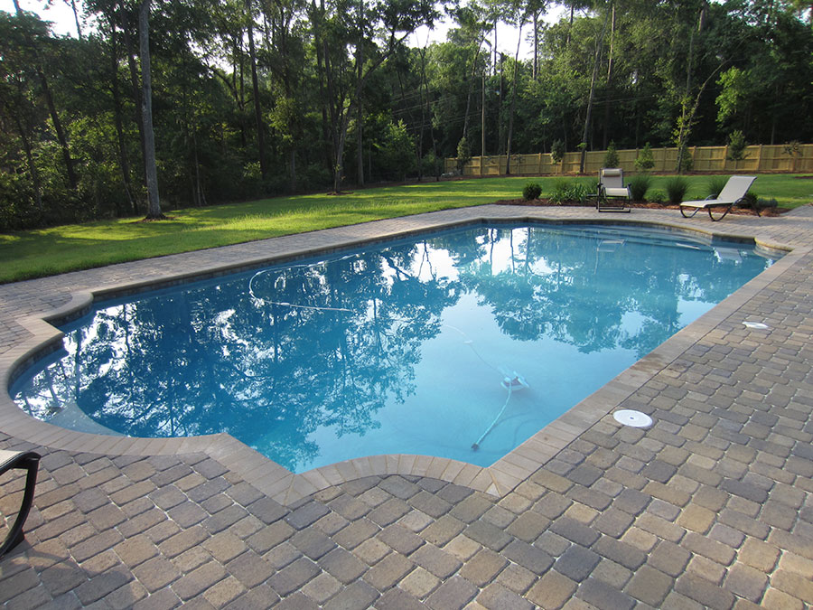 Mystic Pools custom saltwater gunite pool in Valdosta with classic Roman-style edge, brick paver decking, lounge chairs, and a wood privacy fence surrounded by wooded landscaping