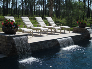 Mystic Pools custom gunite pool with dual sheer waterfall features, stone coping, white lounge chairs, and decorative flower planters