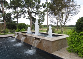 Mystic Pools custom raised pool feature with three vertical fountain jets and spillover waterfall into lower pool, surrounded by mature landscaping