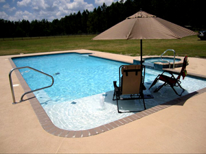 Mystic Pools custom rectangular gunite pool with attached spa, concrete pool deck, patio chairs, and umbrella in an open backyard setting