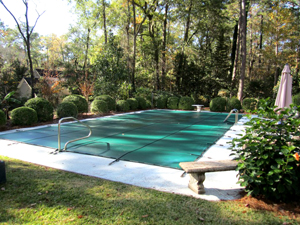 Mystic Pools custom rectangular gunite pool with green safety cover installed, surrounded by manicured shrubs and wooded backyard landscaping